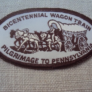 Könnte beinhalten: Ovaler Aufnäher mit braunem Rand und hellblauem Zentrum. Der Aufnäher zeigt eine braune Illustration eines Planwagens, der von Pferden gezogen wird, mit dem Text "BICENTENNIAL WAGON TRAIN PILGRIMAGE TO PENNSYLVANIA".