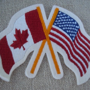 May include: Embroidered patch featuring the flags of Canada and the United States crossed. The Canadian flag has a red maple leaf on a white background with red bars. The American flag has red and white stripes with blue stars.