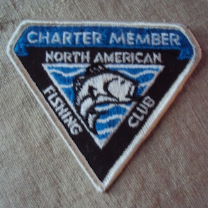Op de afbeelding: Een witte, zwarte en blauwe driehoekige patch met de tekst "Charter Member North American Fishing Club".