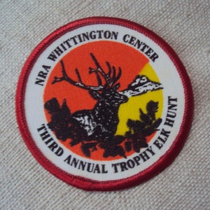 May include: A white, red, and yellow circular patch with a red border. The patch features a black and white illustration of an elk with large antlers. The text "NRA Whittington Center Third Annual Trophy Elk Hunt" is printed around the edge of the patch.