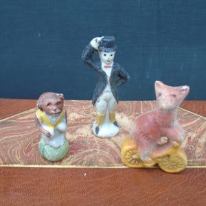 May include: Three small, vintage ceramic figurines. A monkey wearing a yellow shawl, a man in a tuxedo and top hat, and a fox riding a yellow bicycle. The figurines are painted in muted colors and appear to be antique.