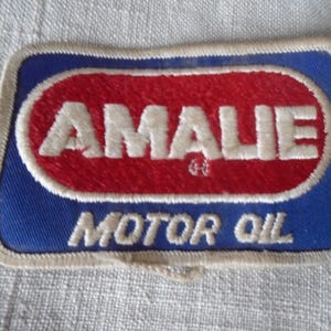 Op de afbeelding: Geborduurde patch met de tekst "AMALIE MOTOR OIL". De patch heeft een blauwe achtergrond met een rode ovaal in het midden. De tekst is wit, met een crèmekleurige rand.
