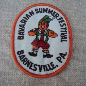 Puede incluir: Un parche ovalado blanco con un borde naranja. El parche presenta a un hombre de dibujos animados con ropa tradicional bávara, sosteniendo una jarra de cerveza. El texto "Bavarian Summer Festival Barnesville, PA" está bordado alrededor del hombre.