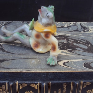 Op de afbeelding: Een keramiek kikker beeldje, geschilderd in groen, geel en bruin. De kikker zit op een boek met een marmeren omslag.