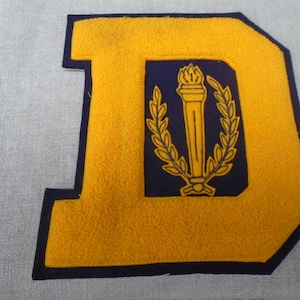 Op de afbeelding: Een gele vilt letter "D" met een marineblauwe rand. Binnen de "D" is een marineblauwe rechthoek met een gouden fakkel en laurierkrans ontwerp. De patch staat op een lichtgrijze stoffen achtergrond.