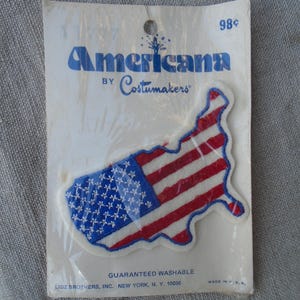 May include: A vintage Americana patch in the shape of the United States, depicting the American flag. The patch is red, white, and blue, with the text "Americana by Costumakers" on the packaging. The packaging also includes the text "Guaranteed Washable".