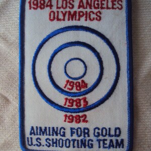 Puede incluir: Un parche blanco con bordado azul y rojo. Presenta un blanco con círculos concéntricos y el texto "1984 LOS ANGELES OLYMPICS" en la parte superior, "1984", "1983" y "1982" dentro del blanco, y "AIMING FOR GOLD U.S. SHOOTING TEAM" en la parte inferior.