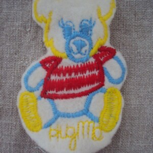 Op de afbeelding: Een witte, geborduurde teddybeer van vilt met gele, blauwe en rode details. De beer draagt een rood-wit gestreept shirt en heeft de woorden "Hug Me" geborduurd op zijn buik.