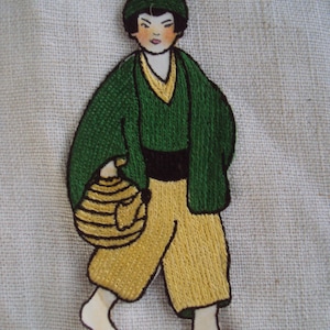 May include: Embroidered image of a person wearing a green jacket and yellow pants, carrying a basket. The person has black hair and a green hat.