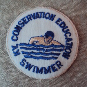 Könnte beinhalten: Ein weißer gestickter Aufnäher mit blauen und braunen Nähten. Der Aufnäher ist rund und zeigt einen Schwimmer in einem Wasserbecken. Der Text um den Schwimmer lautet "KYL CONSERVATION EDUCATION SWIMMER".