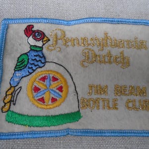 Puede incluir: Parche bordado con fondo gris claro y borde azul. El parche presenta un diseño de ave colorida y las palabras "Pennsylvania Dutch" y "Jim Beam Bottle Club" en letras doradas.