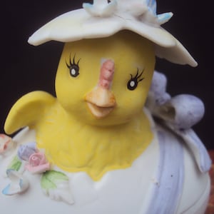 May include: A ceramic figurine of a yellow chick emerging from a white eggshell. The chick has black eyelashes and a pink and orange beak. The eggshell is decorated with pastel flowers and a blue ribbon. The chick is wearing a white hat with pink and blue accents.