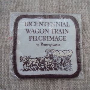 Puede incluir: Un parche bordado marrón y blanco con el texto "Bicentennial Wagon Train Pilgrimage to Pennsylvania". El parche presenta una ilustración en blanco y negro de un carro cubierto tirado por caballos.