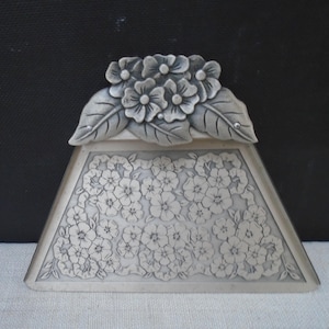 May include: A silver-toned metal dustpan featuring a trapezoidal shape with a flat base. The handle area is decorated with a raised, three-dimensional floral motif of blossoms and leaves, while the main body displays an etched floral pattern.