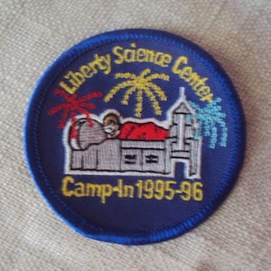 May include: Blue and yellow embroidered patch with the text "Liberty Science Center Camp-In 1995-96". The patch features a cartoon illustration of a person sleeping in a bed with fireworks in the background.