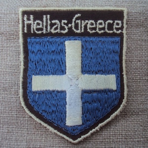 May include: Embroidered patch with a brown border and a blue background featuring a white cross. The text "Hellas-Greece" is embroidered in white on the top of the patch.