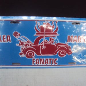 以下が含まれることがあります： 赤いイラストが描かれた青い金属製のナンバープレート。車には物が積まれています。「FLEA MARKET FANATIC」の文字がプレートに印刷されています。