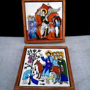 May include: Two square ceramic tiles with religious illustrations, set in wooden frames. One depicts an angel and figures near a tomb, the other shows a figure on a donkey with others. The tiles feature vibrant colors and detailed artwork.