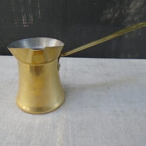 May include: A polished brass Turkish coffee pot featuring a wide, flared base, a tapered body, and a long, slender handle attached with a single rivet. The interior of the pot is lined with a silver-toned metal, and the rim includes a pouring spout.