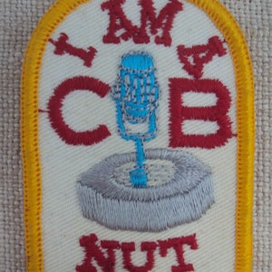 Könnte beinhalten: Ein weißer gestickter Aufnäher mit gelbem Rand. Der Aufnäher zeigt ein rotes Mikrofon mit blauer Spitze, einen grauen Kreis und den Text "I AM A CB NUT".
