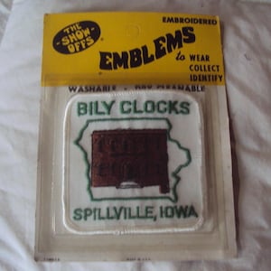 May include: White embroidered patch with green trim. The patch features a brown rectangular sign with the text "Bily Clocks" and the text "Spillville, Iowa". The patch is packaged in a clear plastic bag with a yellow header that reads "The Show-Offs Embroidered Emblems to Wear Collect Identify".