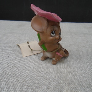 May include: A small brown ceramic mouse figurine with large ears and painted facial features. The mouse holds a large pink flower over its head with a green stem. A small beige paper tag is attached to the figurine with a thin string.