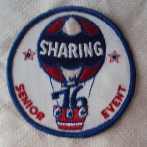 Puede incluir: Un parche ovalado blanco con costuras azules alrededor de los bordes. Un globo aerostático rojo, blanco y azul con la palabra "SHARING" en bordado azul en el lateral. El globo vuela sobre el número "76" en bordado azul. Las palabras "SENIOR EVENT" están bordadas en rojo en la parte inferior del parche.
