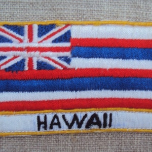 May include: Embroidered patch featuring the Hawaii state flag. The design includes the Union Jack in the upper left corner, with alternating red, white, and blue stripes. The word "HAWAII" is embroidered in white letters at the bottom, with a yellow border.