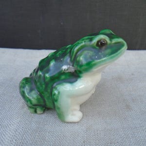 May include: A ceramic frog figurine with a glossy green and white finish. The frog is in a sitting position, with its legs tucked beneath its body. The figurine has detailed features, including eyes and textured skin.