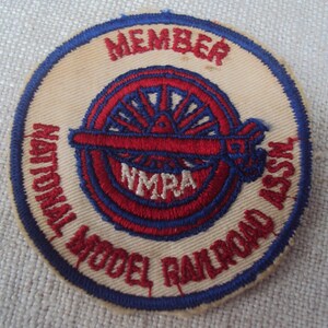 May include: A white, blue and red embroidered patch with the text "MEMBER NATIONAL MODEL RAILROAD ASSN." and "NMRA" in the centre.