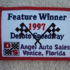 May include: White embroidered patch with red border. It says "Feature Winner 1997 DeSoto Speedway Angel Auto Sales Venice, Florida". A checkered flag is embroidered on each side of the text. A smaller checkered flag with the letters "DS" is in the lower left corner.