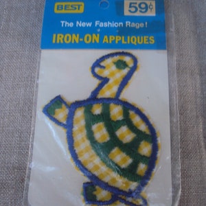 Op de afbeelding: Een strijk-op applicatie van een schildpad met een ruitjespatroon, in blauw en geel. De verpakking vermeldt "The New Fashion Rage! IRON-ON APPLIQUES", "BEST" en "59¢".