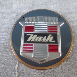 Può includere: Un emblema per auto Nash vintage. L'emblema è circolare con uno sfondo blu scuro e un bordo color oro. Il design centrale presenta uno scudo argentato con accenti rossi e argentati e il nome "Nash" in un rettangolo nero.