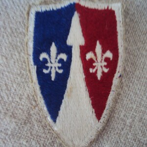 Könnte beinhalten: Ein weißer, blauer und roter gestickter Aufnäher in Form eines Schildes mit einer Fleur-de-lis auf jeder Seite.