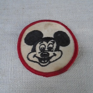 Op de afbeelding: Een ronde geborduurde patch met een zwart-witte illustratie van het personage Mickey Mouse. Het ontwerp is gecentreerd op een gebroken witte stoffen achtergrond en is omlijst door een dikke, felrode gestikte rand langs de buitenkant.