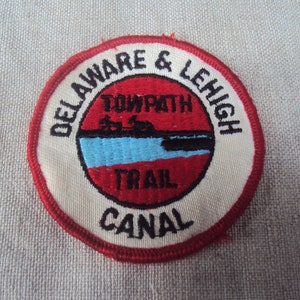 May include: A round patch with a red border and a cream background. The patch has the text "DELAWARE & LEHIGH" and "TOWPATH TRAIL CANAL" in black. A red circle in the centre features a blue river scene and black silhouettes.