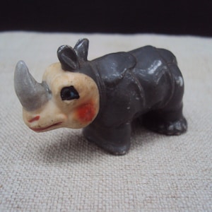 Puede incluir: Una pequeña figura de rinoceronte de cerámica gris y blanca con la cara pintada. La figura tiene un cuerno detallado y un cuerpo texturizado.