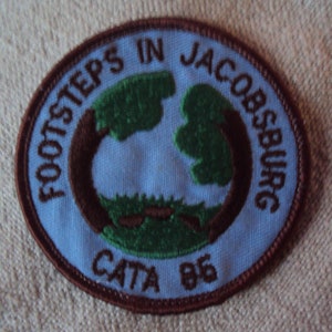 Puede incluir: Parche bordado marrón con fondo azul. El parche dice "Footsteps in Jacobsburg CATA '85" con un diseño verde y marrón en el centro.