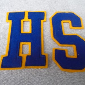 May include: Two felt patches, each featuring a letter in a varsity style. The letter "H" and the letter "S" are in a deep blue color with a gold border. The patches are likely for a high school or sports team.