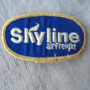 May include: Embroidered patch with the text "Skyline airfreight" in white on a blue oval. The patch has a gold border and is set against a light blue fabric background.