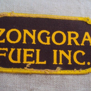 Puede incluir: Un parche ovalado marrón con costuras amarillas alrededor de los bordes. El texto "ZONGORA FUEL INC." está impreso en amarillo en el parche.