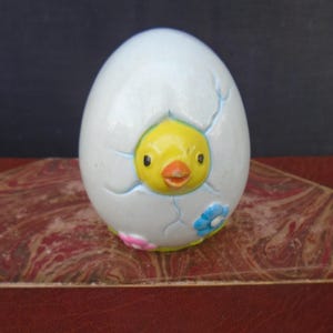 May include: A ceramic Easter egg with a chick hatching from it. The egg is white with blue cracks and small pink and blue flowers. The chick has a yellow head, orange beak, and black eyes. The egg is sitting on a red and brown surface.