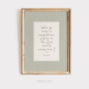 Puede incluir: Arte de pared enmarcado con un marco de madera clara. La obra de arte presenta un fondo color crema con un patrón de rayas verticales en verde claro. El texto dice: "Cuando mi corazón está abrumado, llévame a la Roca que es más alta que yo."