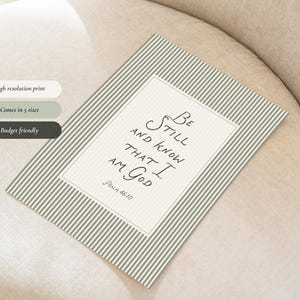 May include: A rectangular print with a striped background in shades of green and white. The print features the text "Be Still and Know That I Am God" in elegant script. The print is displayed on a neutral-toned surface.