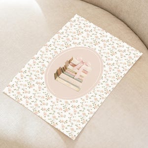 May include: A floral print card featuring a stack of books tied with a pink ribbon, set within an oval frame. The card has a white background with small pink flowers and green leaves. The image is on a beige surface.