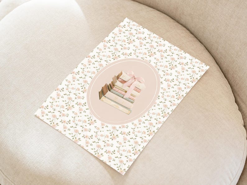 May include: A floral print card featuring a stack of books tied with a pink ribbon, set within an oval frame. The card has a white background with small pink flowers and green leaves. The image is on a beige surface.