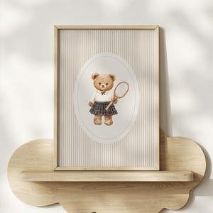 K&ouml;nnte beinhalten: Ein gerahmter Aquarelldruck mit einem Teddyb&auml;ren in einem wei&szlig;en Hemd, einem karierten Rock und einem Tennisschl&auml;ger. Der Hintergrund hat vertikale Streifen in Beige und Wei&szlig;. Der Druck wird auf einem Holzregal ausgestellt.