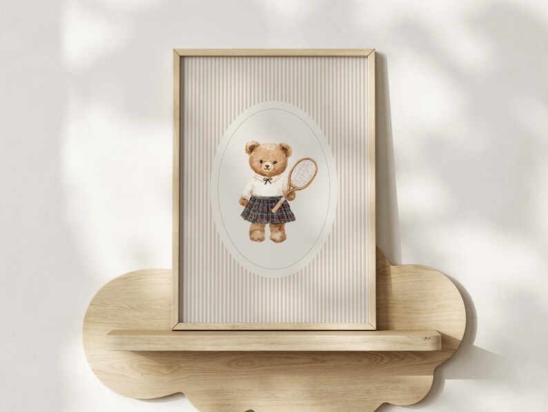K&ouml;nnte beinhalten: Ein gerahmter Aquarelldruck mit einem Teddyb&auml;ren in einem wei&szlig;en Hemd, einem karierten Rock und einem Tennisschl&auml;ger. Der Hintergrund hat vertikale Streifen in Beige und Wei&szlig;. Der Druck wird auf einem Holzregal ausgestellt.