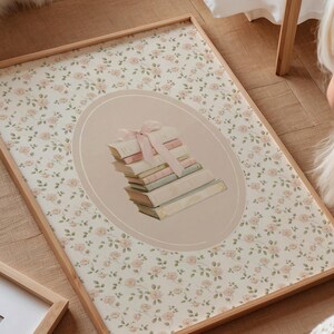 May include: A framed print featuring a stack of books tied with a pink ribbon, set within an oval on a floral background. The frame is light wood. The background has a pattern of small pink roses.