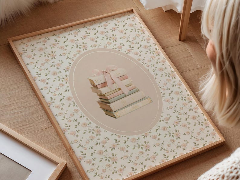 May include: A framed print featuring a stack of books tied with a pink ribbon, set within an oval on a floral background. The frame is light wood. The background has a pattern of small pink roses.